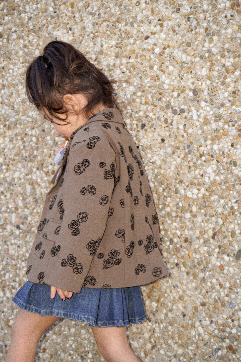 Child wearing a patterned brown jacket and denim skirt on a pebbled surface